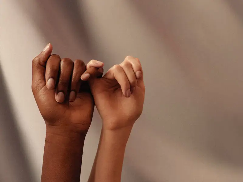 Zwei Hände unterschiedlicher Hautfarbe sind vor einem unscharfen, einfarbigen Hintergrund zu sehen. Die Hände sind so positioniert, dass die kleinen Finger ineinander verhakt sind. Die linke Hand hat eine dunkle Hautfarbe, die rechte Hand eine helle Hautfarbe. Beide Hände sind entspannt und zeigen keine weiteren Accessoires oder Schmuck.
