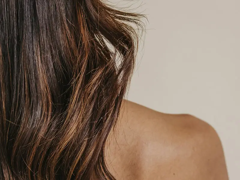 Nahaufnahme eines nackten Rückens und einer Schulter einer Person mit langen, braunen Haaren, die leicht gewellt sind. Die Haare fallen locker über die Schulter, der Hintergrund ist einfarbig hell und unscharf.