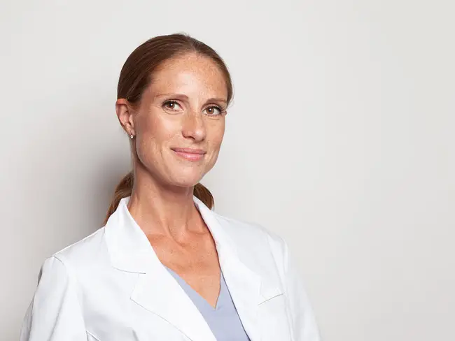 Hautärztin in weißem Arztkittel und hellblauem Oberteil vor neutralem Hintergrund