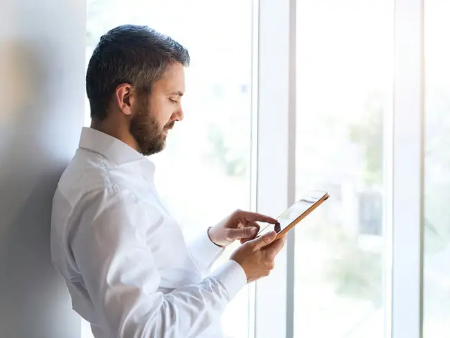 Praxismanager in weißem Hemd steht seitlich vor einem Fenster und bedient ein Tablet mit einer Hand.