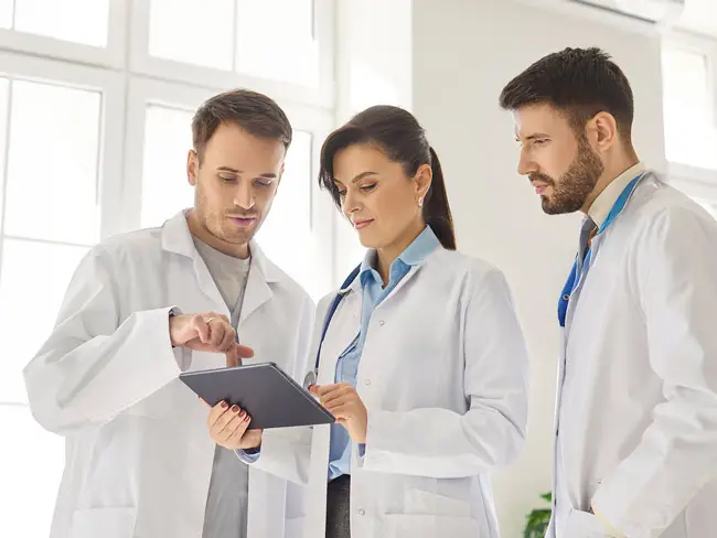 Drei Personen in weißen Arztkitteln stehen in einem hellen Raum mit großen Fenstern. Sie betrachten gemeinsam ein Tablet, das von der Person in der Mitte gehalten wird. Die Person links zeigt mit dem Finger auf das Tablet, während die Person rechts mit verschränkten Armen zuhört. Alle tragen Hemden unter den Kitteln, die Person in der Mitte hat ein hellblaues Hemd an. Im Hintergrund ist eine grüne Pflanze zu erkennen.