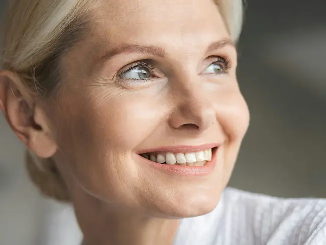 Frau mit blondem Haar hält ein kleines, rundes Spiegelglas vor ihr Gesicht in einer Hautarztpraxis.