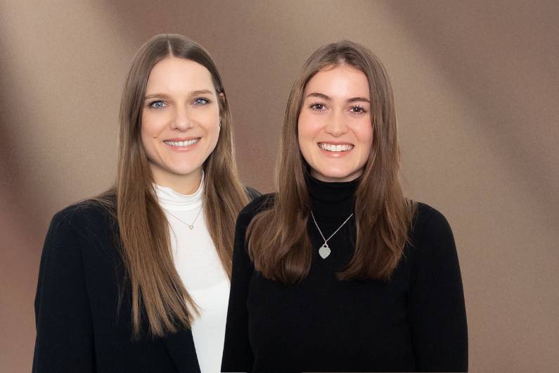 Unsere HR-Abteilung Frau Hellhuber in schwarzem Pullover mit Schmuckanhänger und Frau Meinicke in weißem Oberteil und schwarzem Blazer, vor einfarbigem Hintergrund.