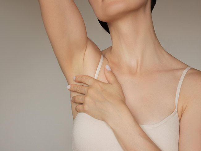 Nahaufnahme einer Frau, die ihren Arm hebt und mit der Hand die Achsel berührt, vor neutralem Hintergrund